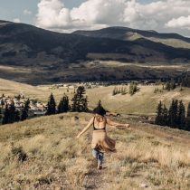 A woman in a tan dress walks down a grassy hill with her arms outstretched, overlooking a small town and forested mountains under a partly cloudy sky.