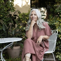 A woman with long gray hair in a mauve dress sits on a white metal chair in a garden beside green plants and a round table, embodying the spirit of Instincts, Intuition & Trusting Yourself (Group).