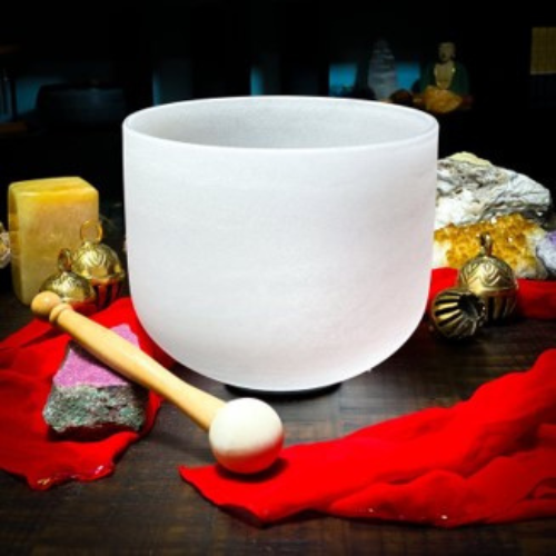 A white crystal singing bowl sits on a wooden table, surrounded by rocks, a candle, gold ornaments, and a red cloth. A mallet rests in front of the bowl, and a small figurine is visible in the background.
