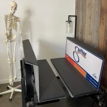 A human skeleton model, used by an injury prevention specialist to demonstrate ACL injury prevention, stands beside a black bench labeled The Shifter Training Session in a white room with a wooden accent wall and lamp.