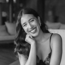 A woman with long wavy hair sits smiling, resting her chin on her hand. She wears a sleeveless top and hoop earrings. The image is in black and white, and the background is softly blurred.