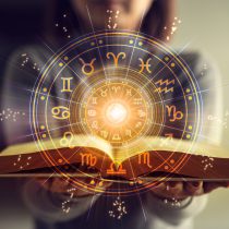 A person holds an open book, with glowing zodiac symbols and a mystical light overlayed above the pages, creating a magical and astrological atmosphere.