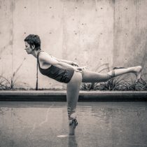 Gabby, a yoga instructor offering a private yoga class, is doing a pose - Warrior 3 - with her standing foot in water.