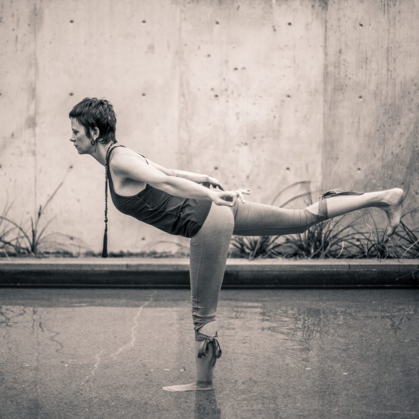 Gabby, a yoga instructor offering a private yoga class, is doing a pose - Warrior 3 - with her standing foot in water.