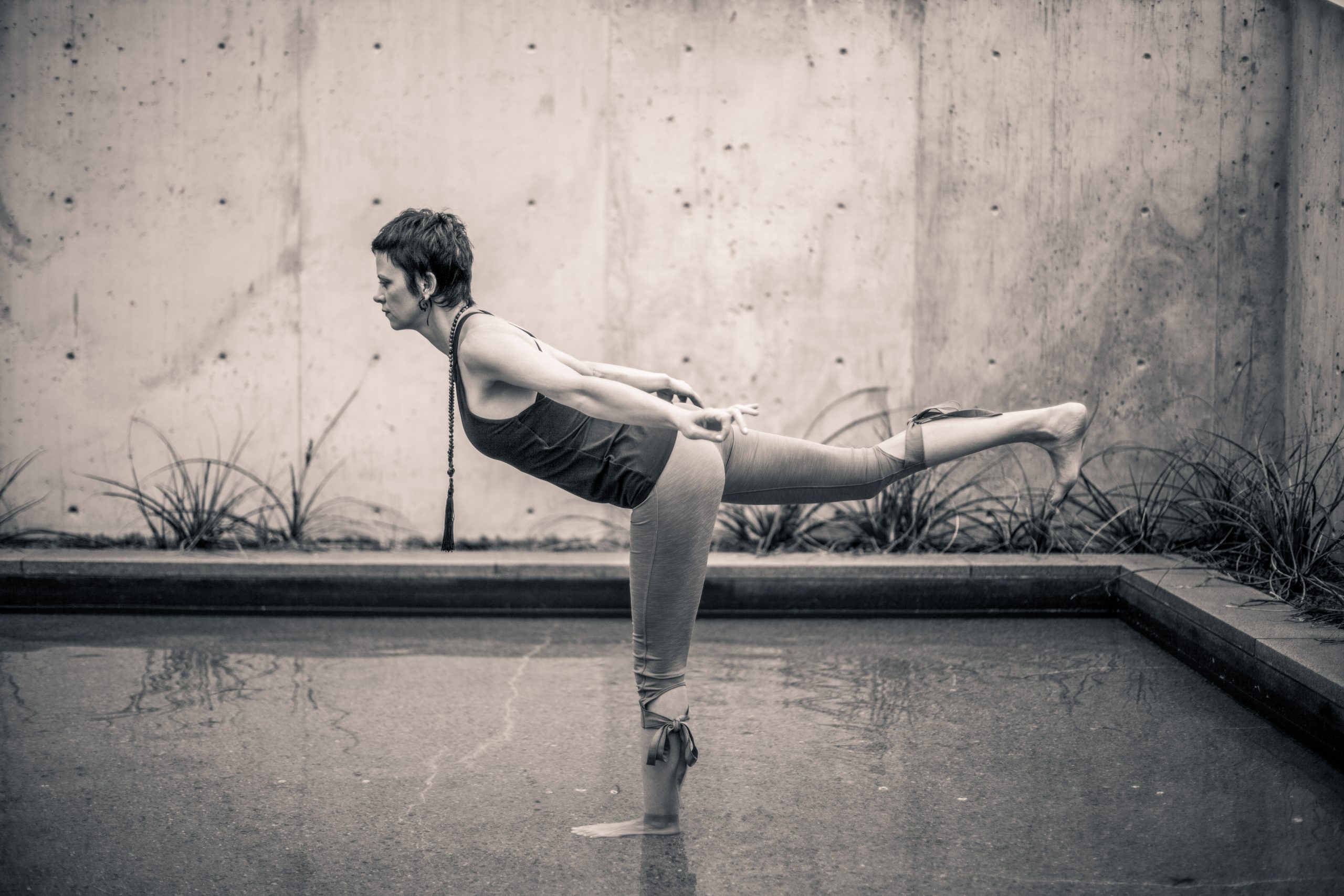 Gabby, a yoga instructor offering a private yoga class, is doing a pose - Warrior 3 - with her standing foot in water.