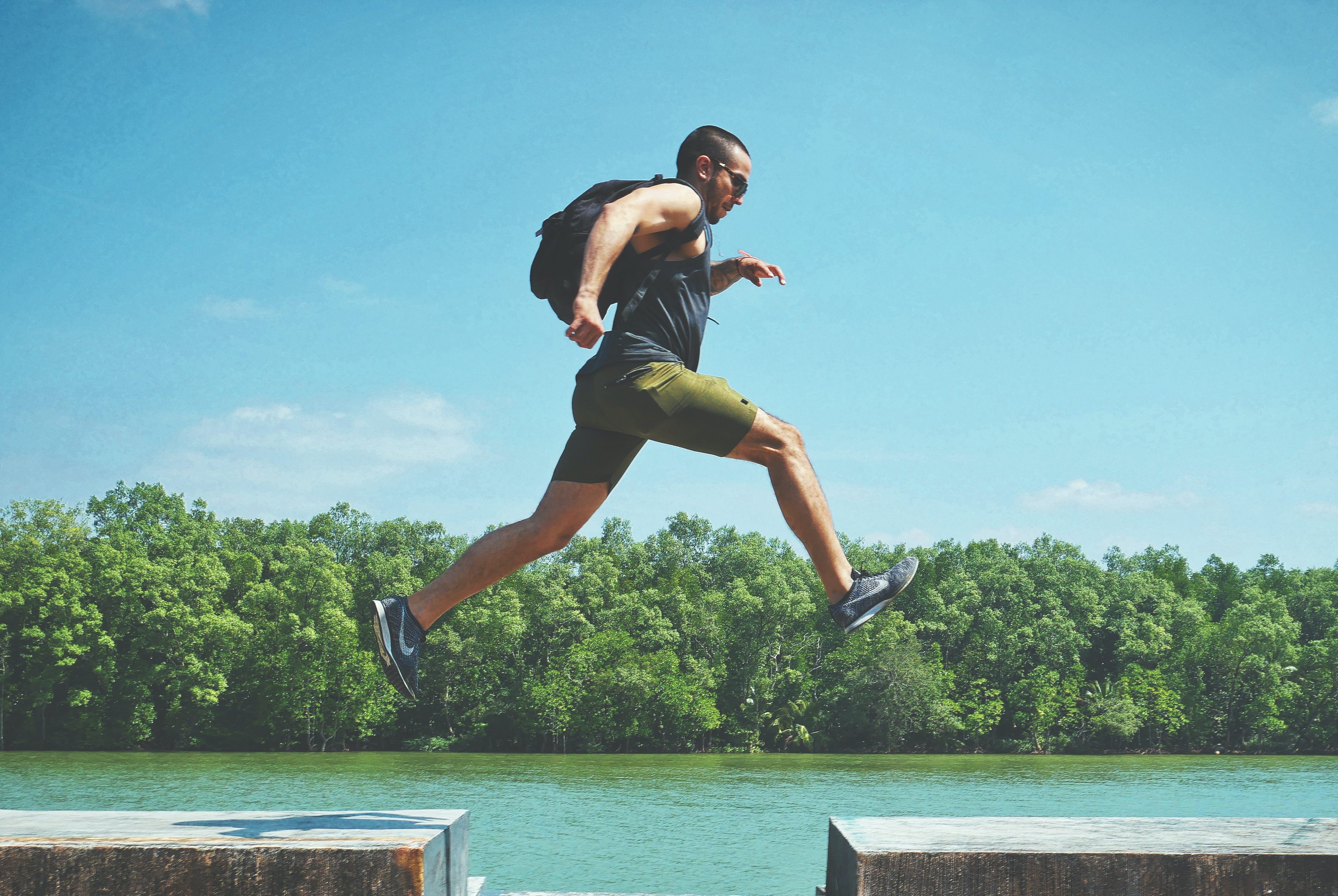 A man leaps between platforms outdoors, wearing athletic clothing and a backpack, with lush trees and a river behind him—embodying the spirit of Develop Your Dreamlife.