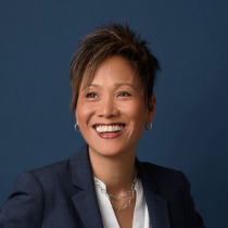 A smiling woman with short, spiky hair in a dark blazer, white blouse, hoop earrings, and a silver chain necklace stands before a dark blue background—your expert for Family and Relationship Consultation.