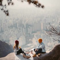 Two people in winter hats share a picnic on a rocky cliff, city skyline below, mist in the air. Pine branches frame this intentional moment from Dating Deliberately.