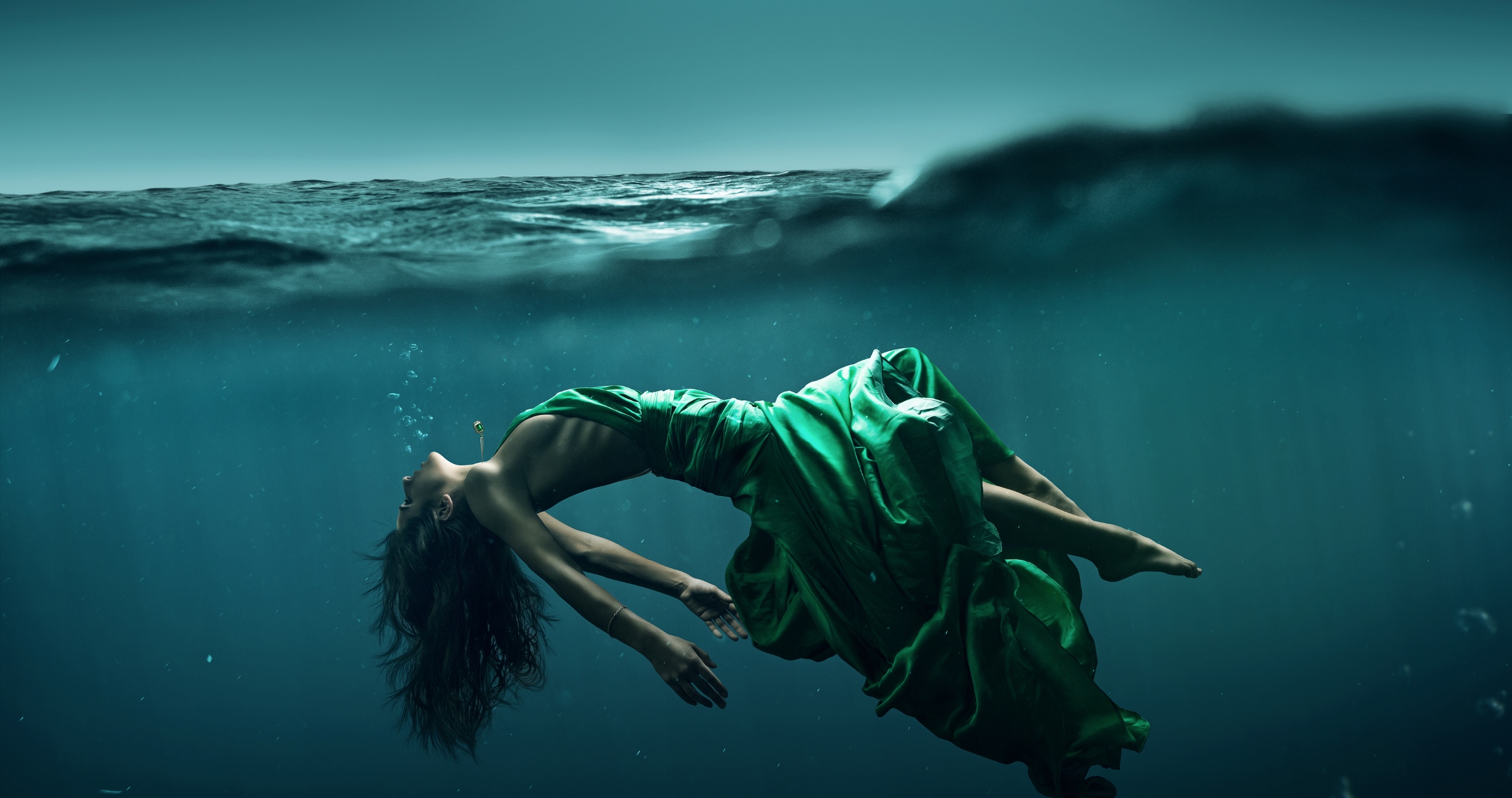 In SHE Breaks Free, a woman in a flowing green dress floats underwater, arching her back and gazing upward as light filters through the surface above.