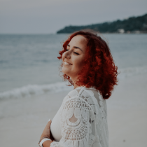 Gerly, who is a somatic awareness coach, is standing on a beach with the ocean behind her smiling. She has curly ready hair and is wearing a white floral shawl.