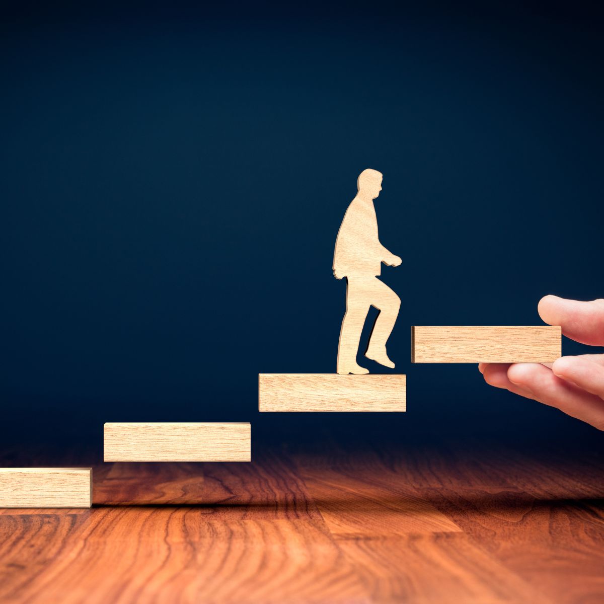 A wooden person climbs a staircase of blocks, with a hand placing the top block, representing progress and support—showcasing Transformative Living Expertise (24) against a dark blue background and wooden surface.
