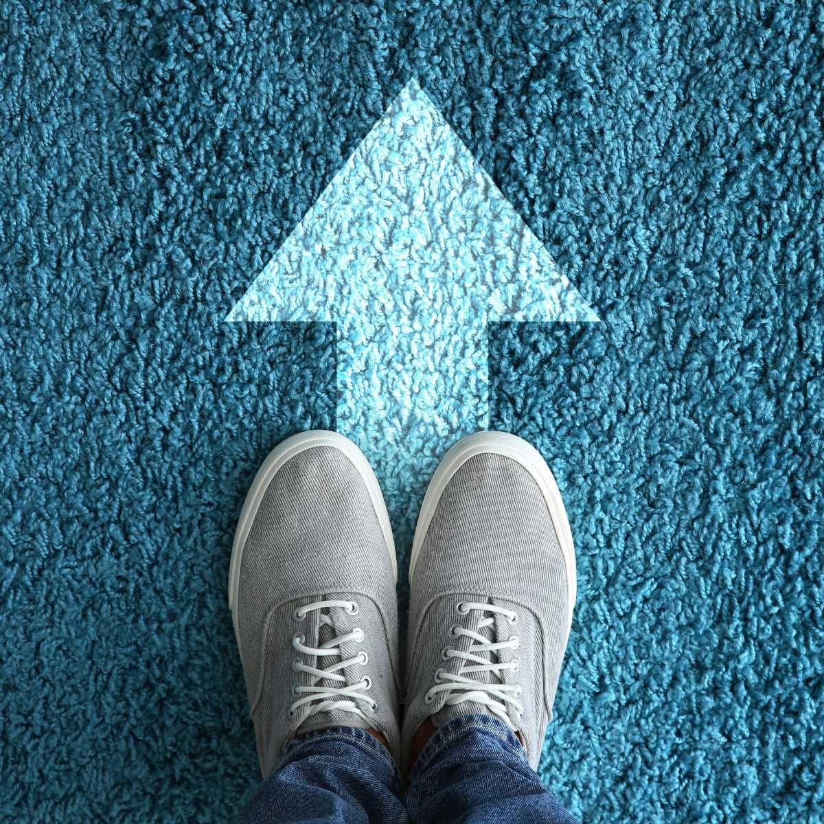 Someone wearing gray sneakers stands on blue carpet facing a large white arrow pointing forward, representing Recovery Expertise (24) and symbolizing the journey toward mental health recovery and new beginnings.