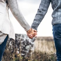 Two people hold hands outdoors, both in cozy sweaters and jeans, embodying the spirit of Dating Deliberately. The blurred grass and rocks suggest a natural autumn or winter setting, perfect for meaningful connections.