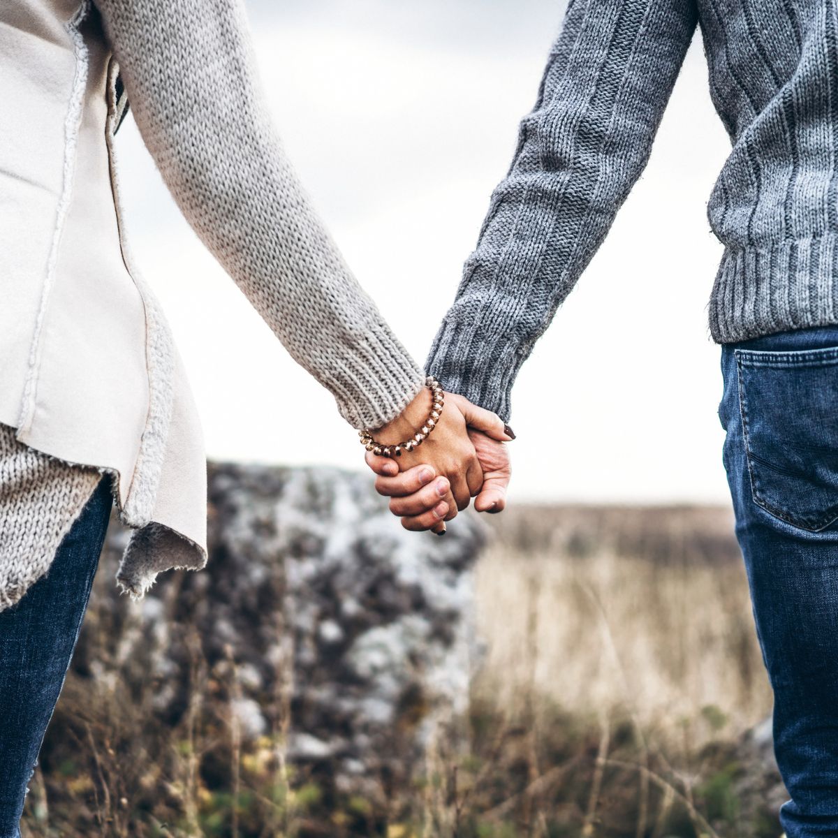 Two people hold hands outdoors, both in cozy sweaters and jeans, embodying the spirit of Dating Deliberately. The blurred grass and rocks suggest a natural autumn or winter setting, perfect for meaningful connections.