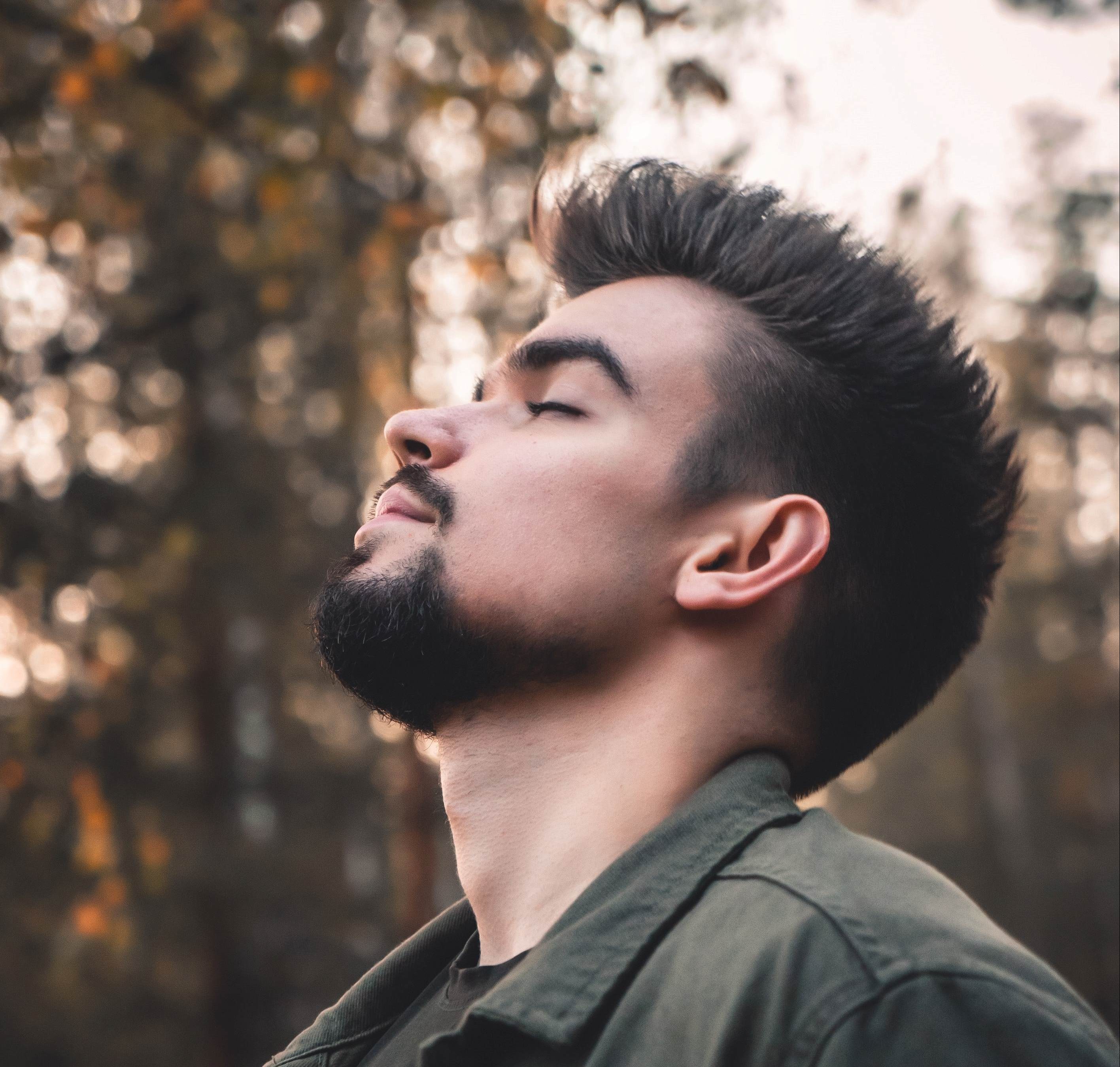 A young man with dark hair and a beard stands outdoors with his eyes closed, looking calm and relaxed, embodying the peace found through the Conscious Breathwork Journey. Sunlight filters through blurred trees in the background.