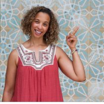 A smiling woman with curly hair wears a red sleeveless top as she points upward, standing before a blue and white geometric wall, promoting the Ageless Grace Seated Brain Health Workout.