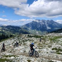 Three cyclists ride mountain bikes along a rocky trail in a scenic mountainous landscape, with green forests and tall peaks under a blue sky with scattered clouds.