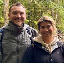 Two people wearing jackets smile together in a forest with green trees and a rocky stream—the perfect setting for Outdoor Wellness Workshops focused on team building activities.