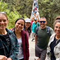 A group of five smiling people pose for a photo on a suspension bridge surrounded by trees during an Outdoor Wellness Workshops event, while others walk along the bridge in the background.
