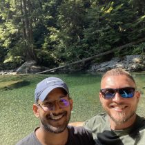 Two men smiling and posing for a selfie in front of a clear river surrounded by rocks and dense green forest on a sunny day.