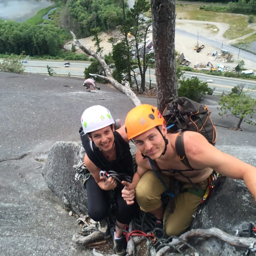 Kevin is pictured here with a client as he helps her build a life of adventure through rock climbing. They are wearing helmets, rock climbing gear, and giving a thumbs up.