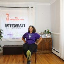 A woman joins the Ageless Grace Seated Brain Health Workout in a bright room with wood floors, exercising on a chair. A banner with The Emotional Tour, INTENSATI, and Ageless Grace logos stands behind her among plants and cabinets.