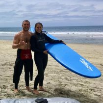 Two people in wetsuits stand smiling on a sandy beach; one holds a large blue surfboard. The ocean and waves are visible in the background under a partly cloudy sky.