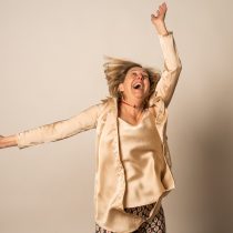 A joyful older woman with blond hair wearing a shiny beige blouse and patterned pants dances with her arms raised, smiling and laughing against a plain light background.