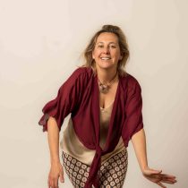 A smiling woman with blonde hair wears a maroon blouse and patterned pants, leaning forward with one arm extended against a plain beige background.