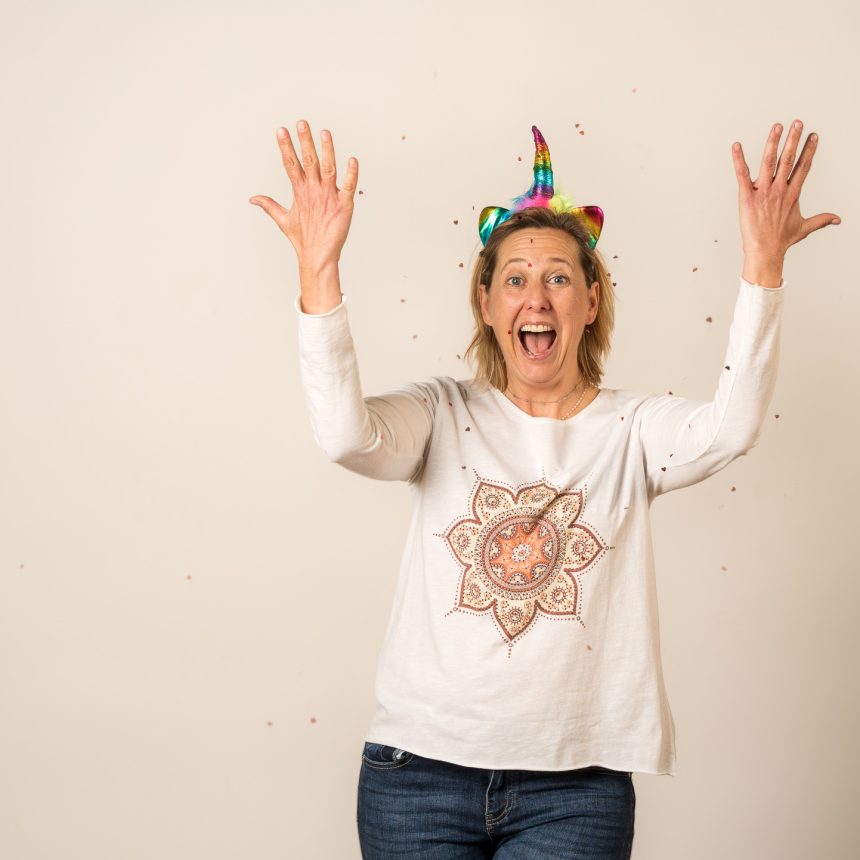 Jenn is wearing a rainbow unicorn headband and white long sleeve shirt with a mandala on it. She has her hands thrown up in the air releasing confetti with her mouth open in joy. This image is to represent her shift your mindset towards joy 30 minute creative boost session.