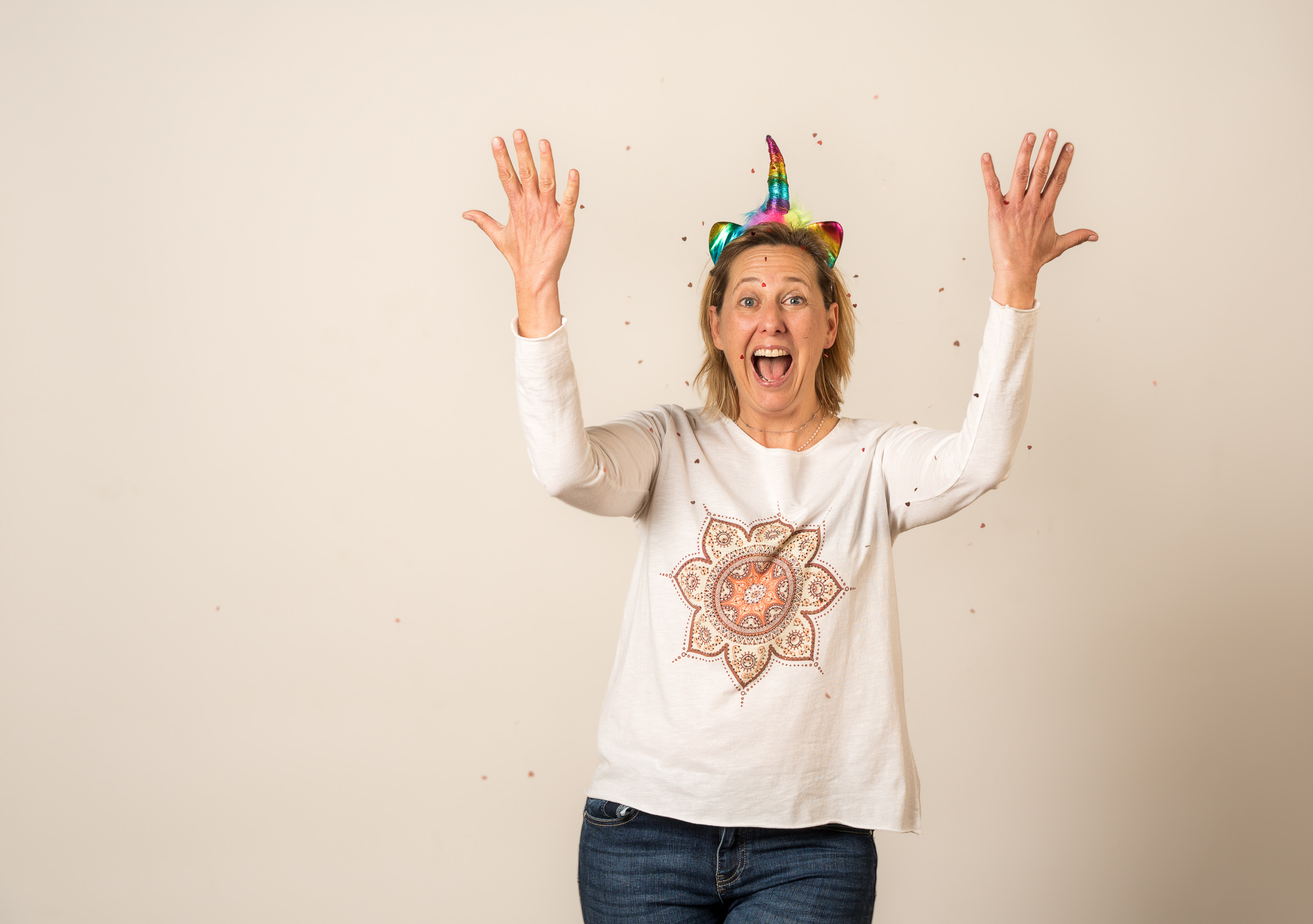 Jenn is wearing a rainbow unicorn headband and white long sleeve shirt with a mandala on it. She has her hands thrown up in the air releasing confetti with her mouth open in joy. This image is to represent her shift your mindset towards joy 30 minute creative boost session.