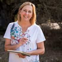 A smiling woman stands outdoors holding a spiral notebook and pen. She wears a white shirt with colorful floral designs and has shoulder-length blonde hair. The background is blurred trees and natural scenery.