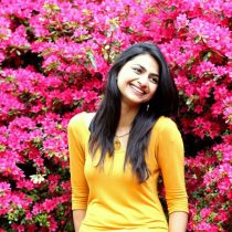 A woman in a yellow top smiles while standing in front of a large shrub filled with vibrant pink flowers.