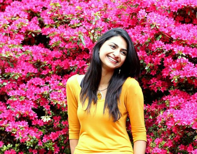 A woman in a yellow top smiles while standing in front of a large shrub filled with vibrant pink flowers.