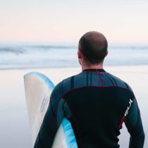A person in a wetsuit stands on a beach holding a surfboard, looking out at the calm ocean under a pale sky.
