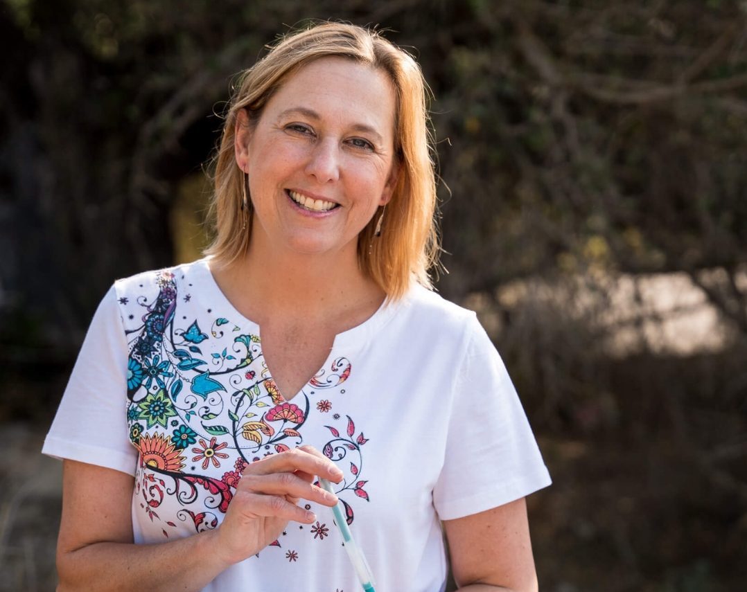 A woman with shoulder-length blonde hair wearing a white shirt with colorful designs smiles while holding a pen and notebook outdoors, with trees and sunlight in the background.