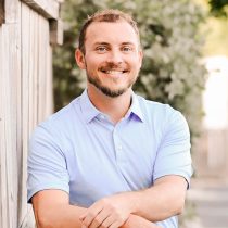 A smiling man with short hair and a beard wears a light blue polo shirt, leaning casually against a wooden fence with greenery in the background.