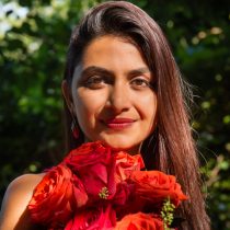 A woman with long brown hair holds a bouquet of red and orange roses, smiling softly. She stands outdoors with green foliage in the background, sunlight highlighting her face.