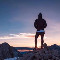 A person enrolled in an adventure program has reached the top of the mountain and is looking at the horizon as the sun rises.