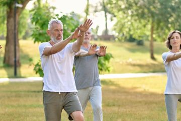 Four adults practice tai chi in a park, standing on grass with arms raised. They wear casual athletic clothing and appear focused, possibly contemplating the difference between chakra and chi as they move. The background includes trees and sunlight.