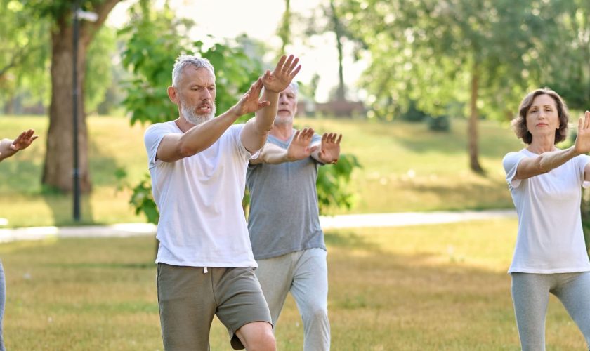 Four adults practice tai chi in a park, standing on grass with arms raised. They wear casual athletic clothing and appear focused, possibly contemplating the difference between chakra and chi as they move. The background includes trees and sunlight.