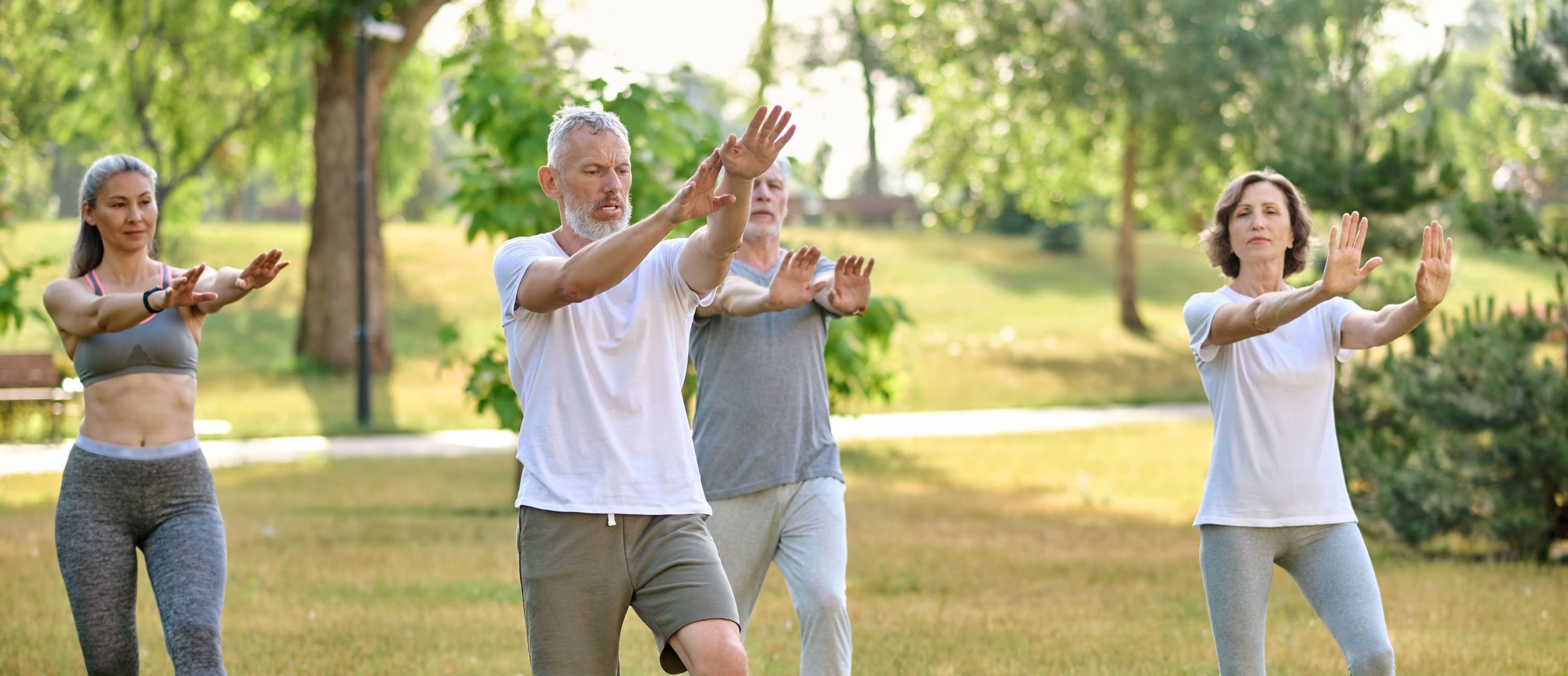 Four adults practice tai chi in a park, standing on grass with arms raised. They wear casual athletic clothing and appear focused, possibly contemplating the difference between chakra and chi as they move. The background includes trees and sunlight.