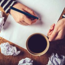 A person enjoying coffee while writing notes amid crumpled papers on a wooden table—capturing the struggle and inspiration behind Cure Your Writers Block.