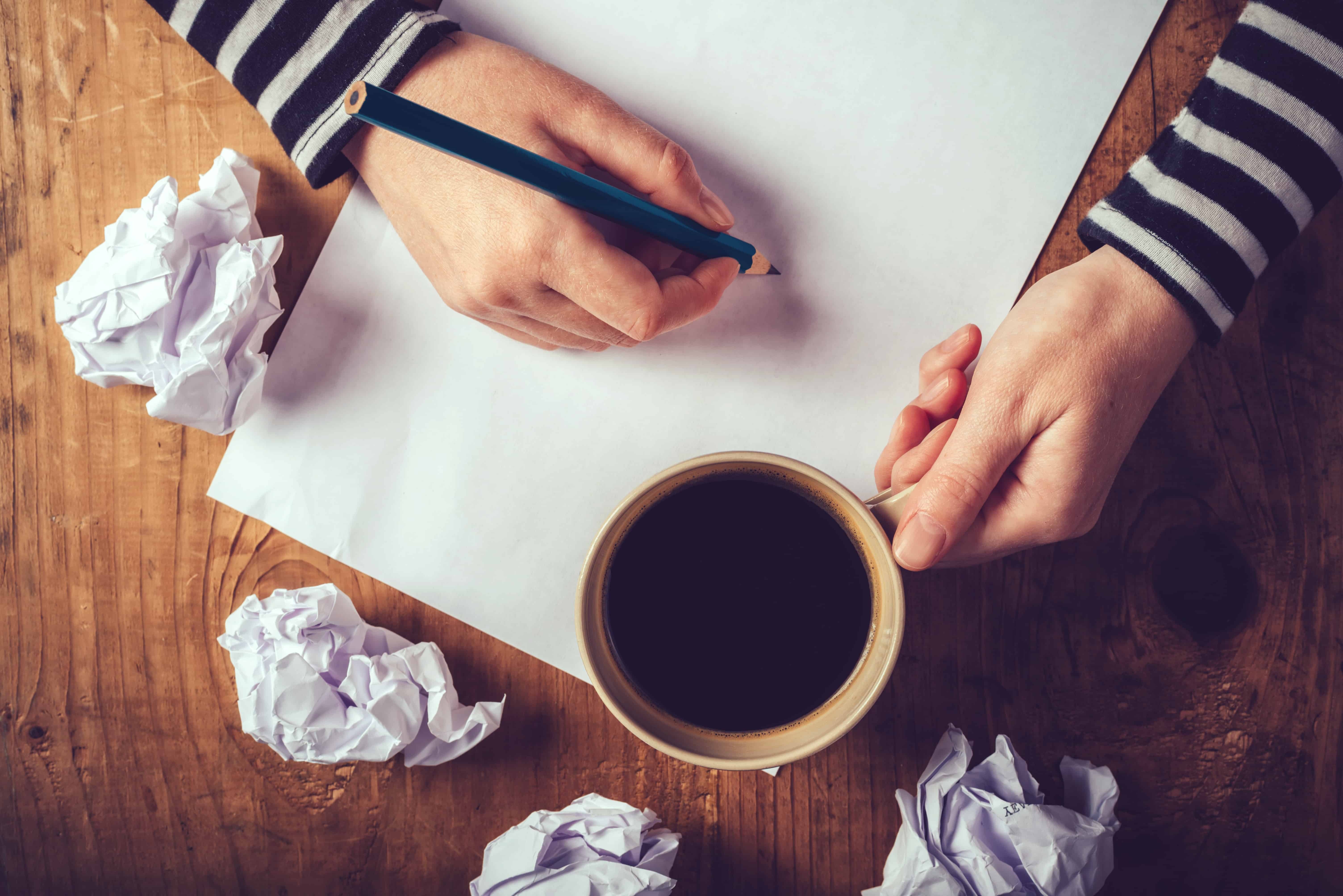 A person enjoying coffee while writing notes amid crumpled papers on a wooden table—capturing the struggle and inspiration behind Cure Your Writers Block.