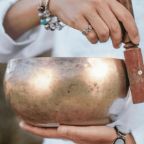 A person uses Sound Healing & Spiritual Guidance, playing a brass singing bowl with a wooden mallet while wearing beaded bracelets, a watch, and a ring. A white sleeve is visible during the private session.