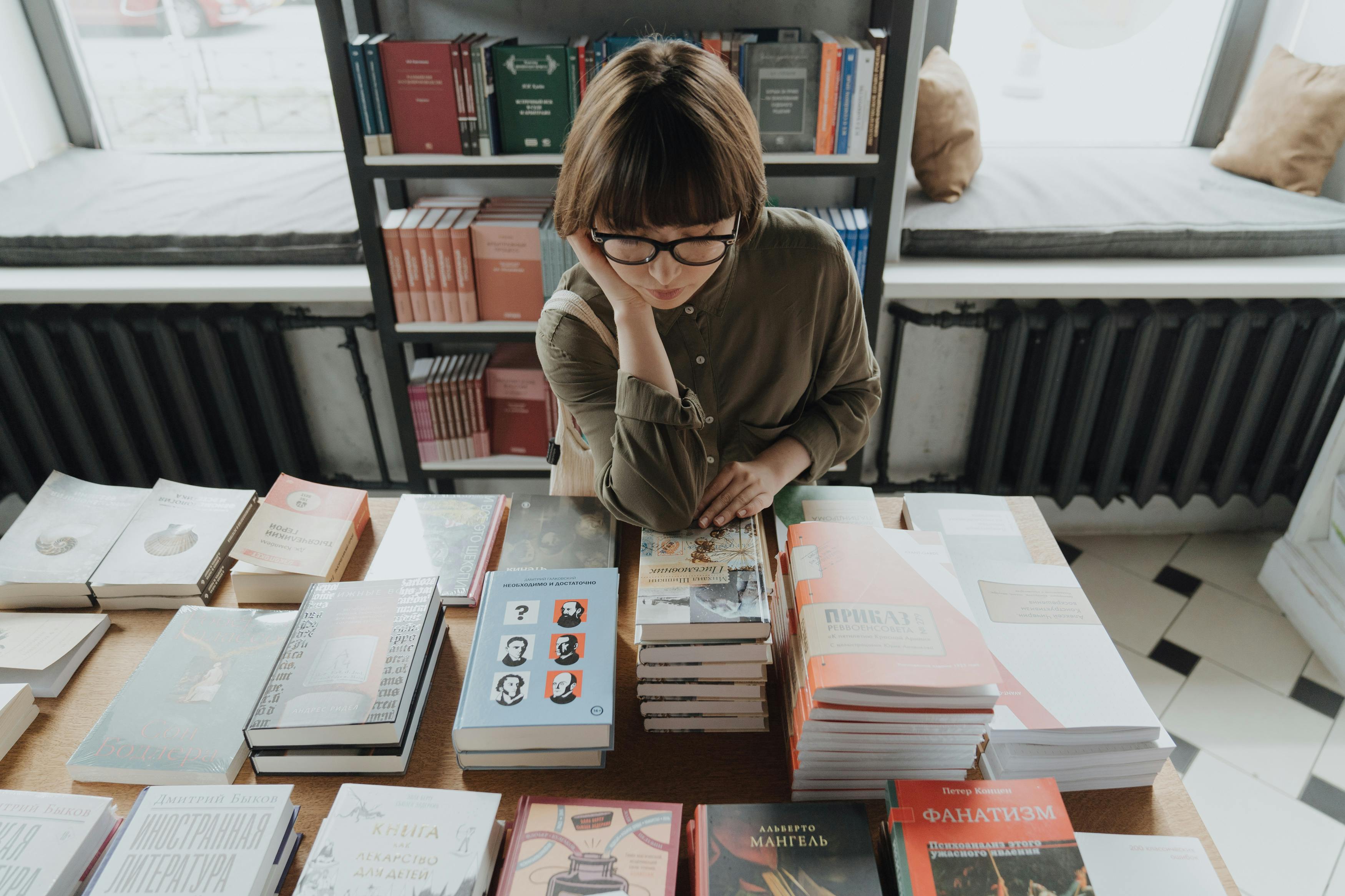 A person with glasses and short hair stands at a table in a bookstore, looking thoughtfully at books, including Sell Your Book: 3-Month Program, while shelves of books line the windowed wall behind them.