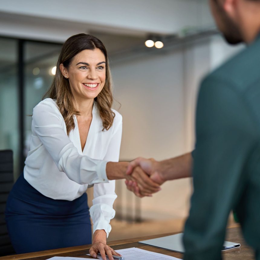 A woman in business attire smiles while shaking hands with someone across a desk in a modern office, exemplifying the confidence gained from the Communicate with Impact & Influence Package.