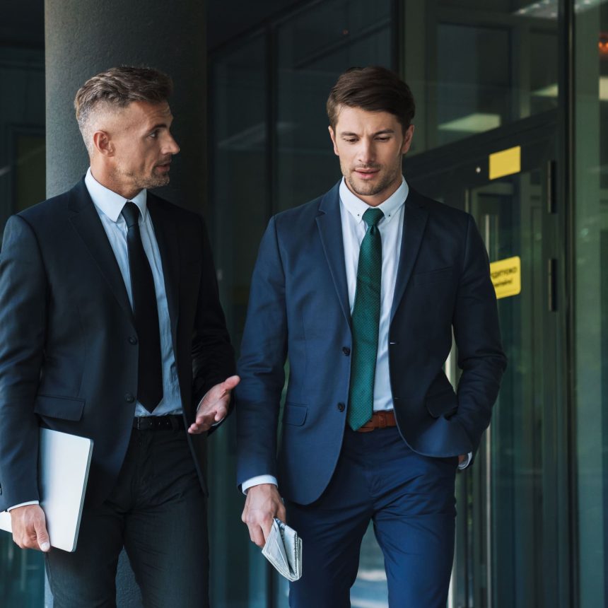 Two men in business suits walk outside a modern glass building; one gestures with a laptop as they discuss the Communicate with Impact & Influence (Package), while the other listens intently, holding a newspaper.