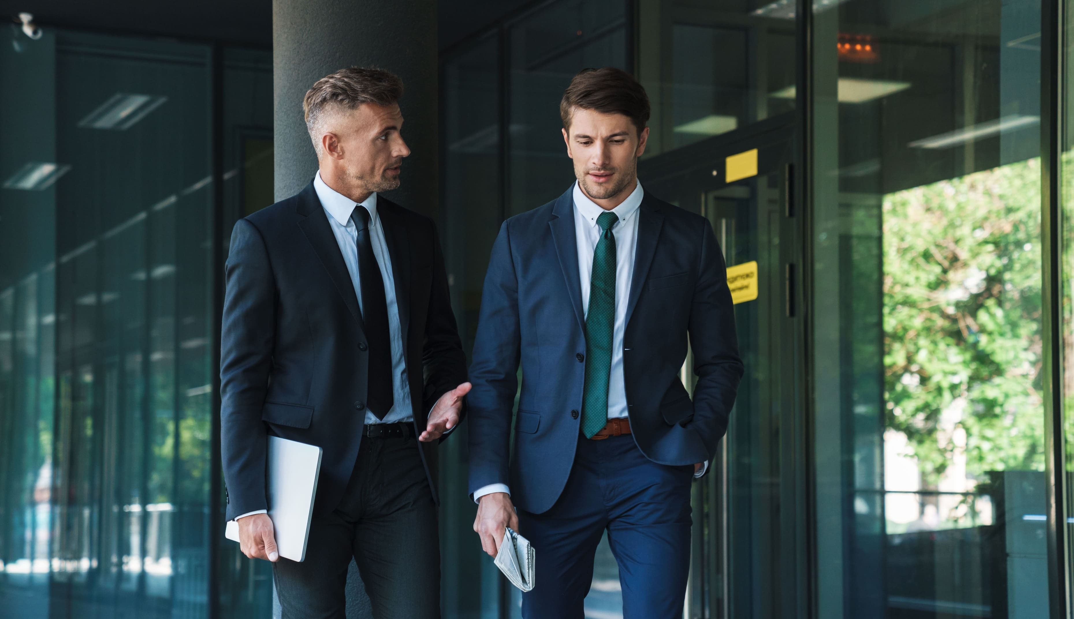 Two men in business suits walk outside a modern glass building; one gestures with a laptop as they discuss the Communicate with Impact & Influence (Package), while the other listens intently, holding a newspaper.