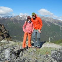 Two people in bright outdoor gear stand on a rocky mountain ledge, smiling together. Its the ideal place for “Communicate with Impact & Influence” coaching, surrounded by snow-capped peaks, forest, and a winding road beneath a clear blue sky.